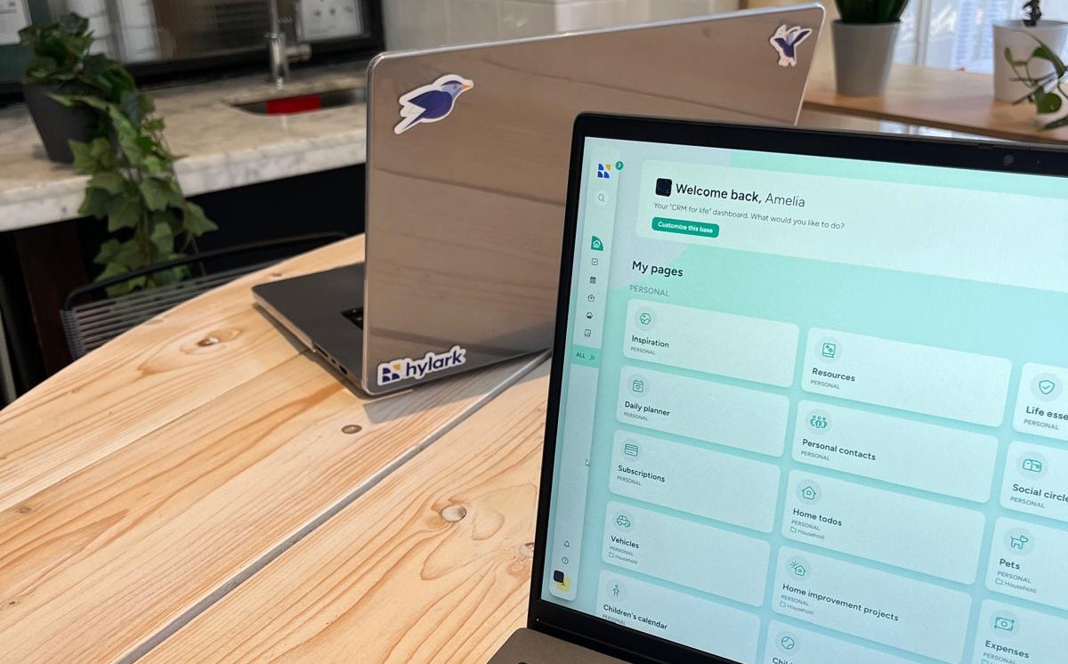 A wooden table with two laptops on it. The closer laptop screen displays a dashboard with the greeting "Welcome back, Amelia" and various sections such as "Inspiration," "Resources," "Daily Planner," and more. The other laptop, positioned further back, has stickers on the lid, including one that reads "Hylark" and another with a bird logo. The background features a countertop with a plant and a kitchen sink.